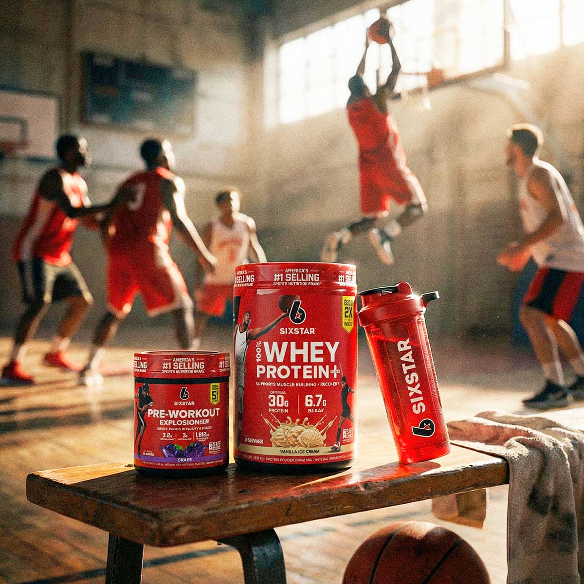 SIXSTAR Performance Starter Stack with whey protein plus, pre-workout explosion 2.0, and shaker cup on gym bench with athletes playing basketball.